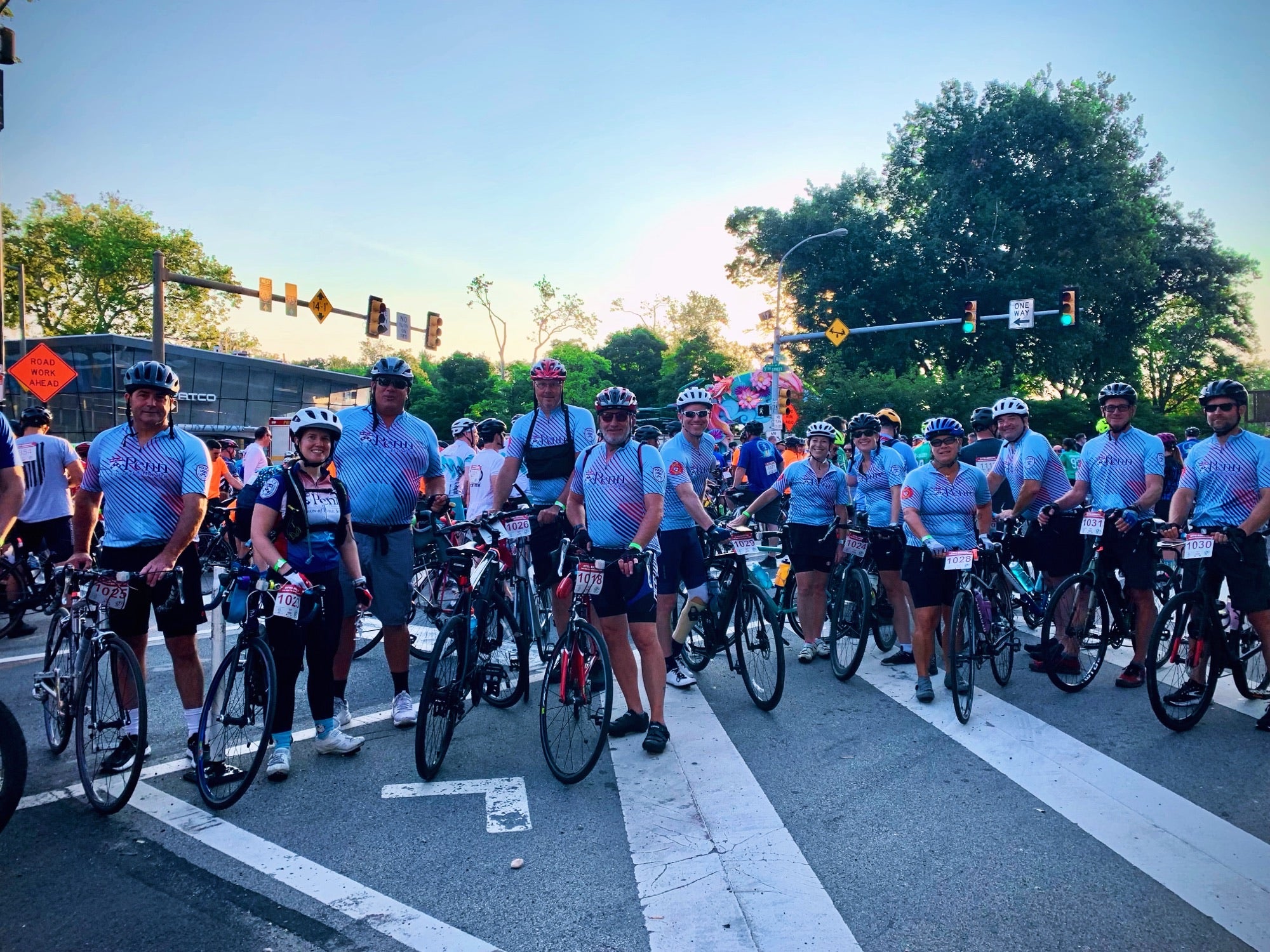 A group of riders wearing matching blue jerseys pose for a photo while on thier bikes. The background is a city street, with the sun rising int he background behind the riders.
