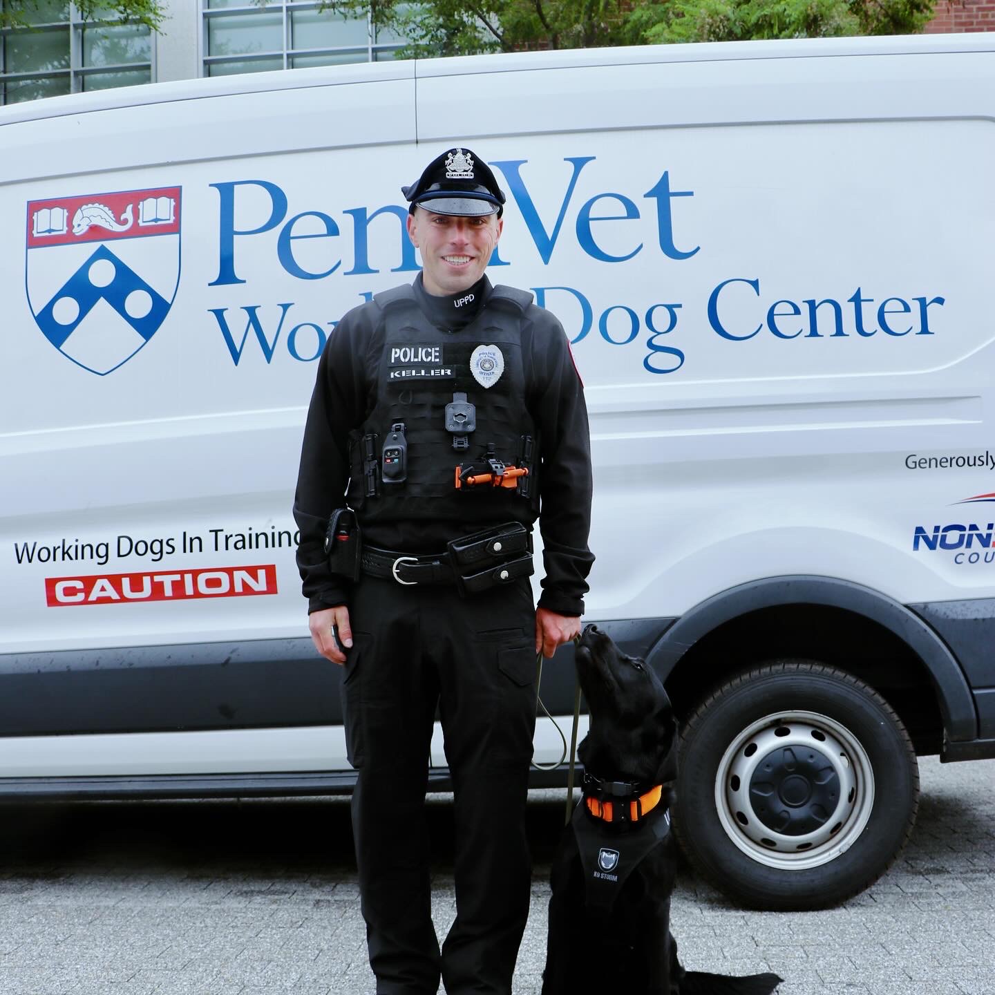 Photo of Office Matthew Keller in a black police uniform standing beside K9 Officer Toby, a black Labrador Retriever, in front of a PennVet Working Dog Center vehicle
