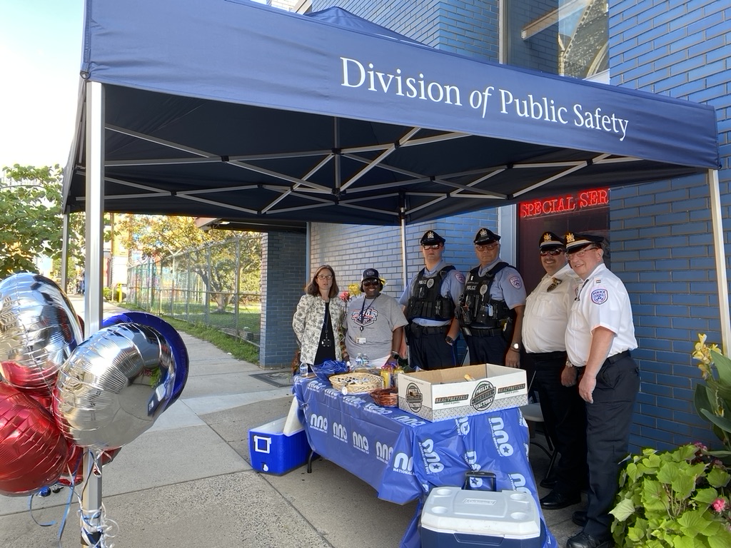 Members of the Division of Public SAfety gather behind a table with pretzels and other giveaway items under a blue tent , in front of the DP HQ, which is a blue brick building.