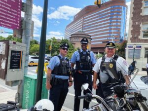 Three police officers pose on a street corner with the Hospital of the University of Pennsylvania in the background