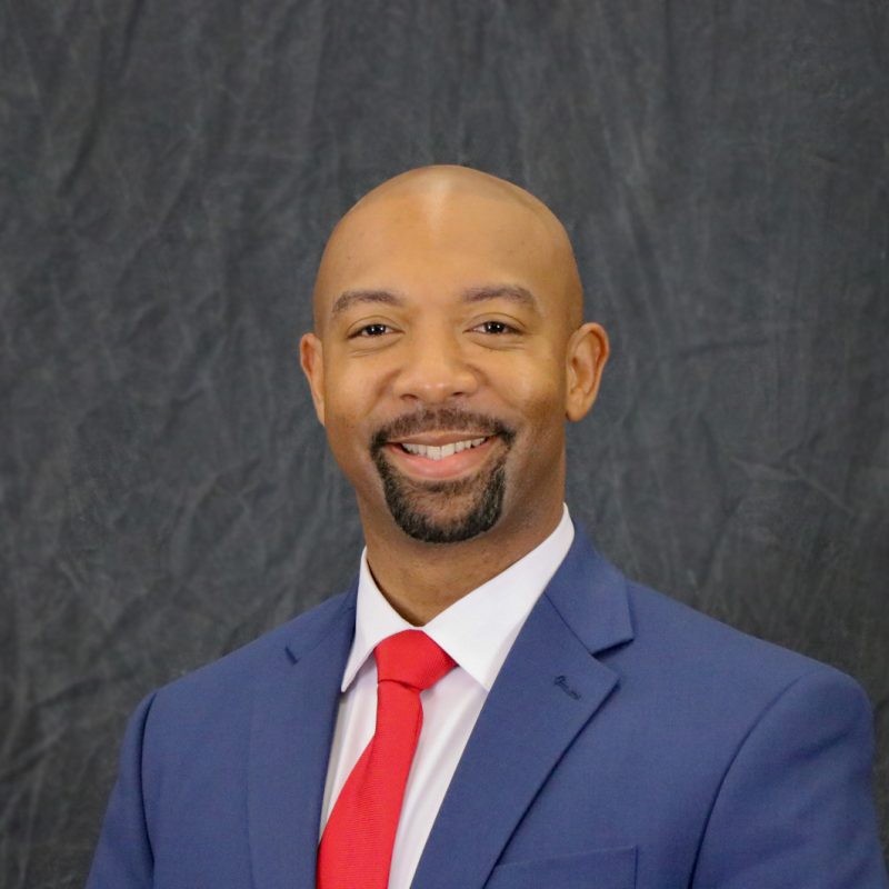 Photo of Josh Forman, short goatee and mustache, having no hair on his head, wearing a blue suit jacket, white shirt, and red tie.