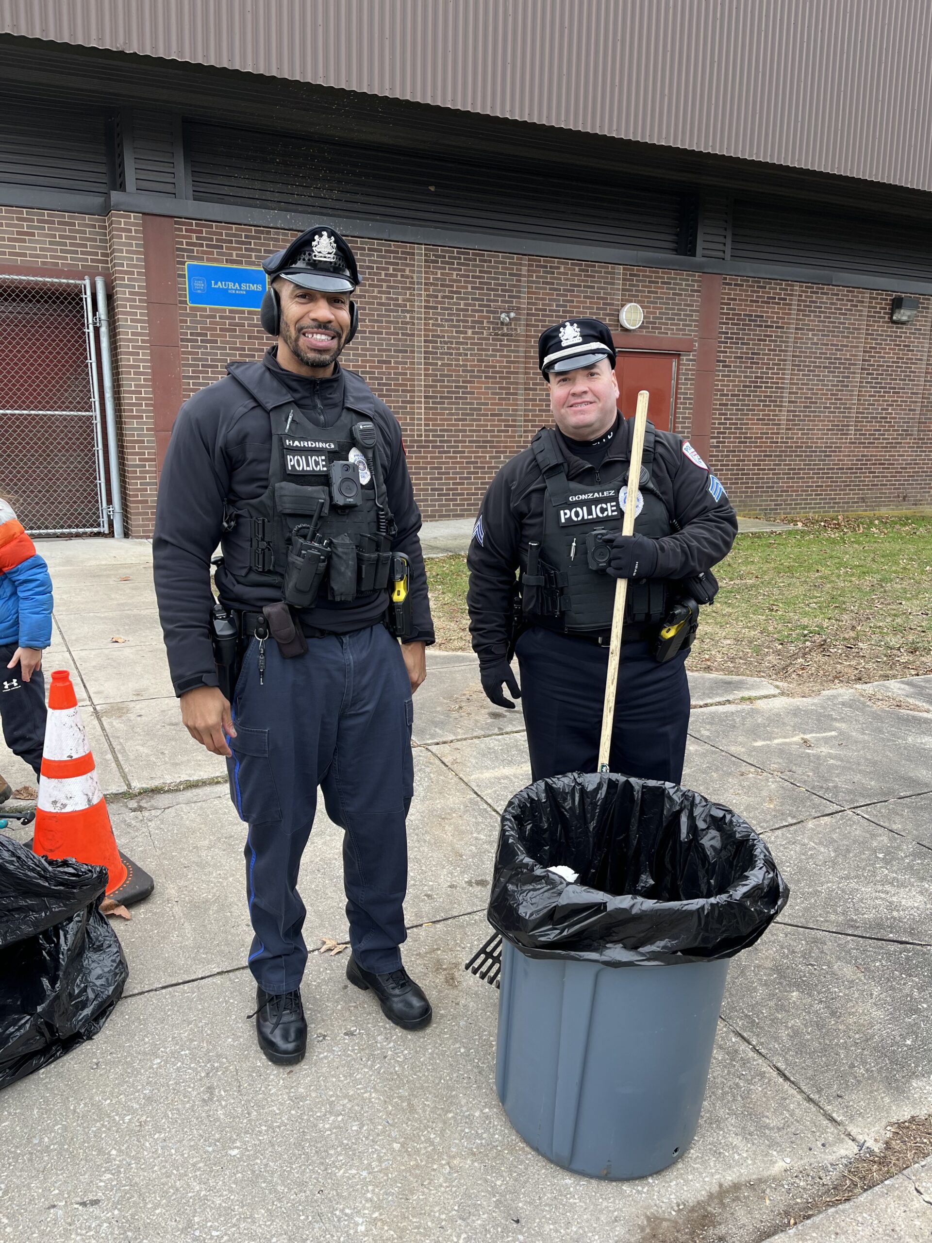 Two officers at a neighborhood clean up event, in front of a brick building with a large trash can and rake.