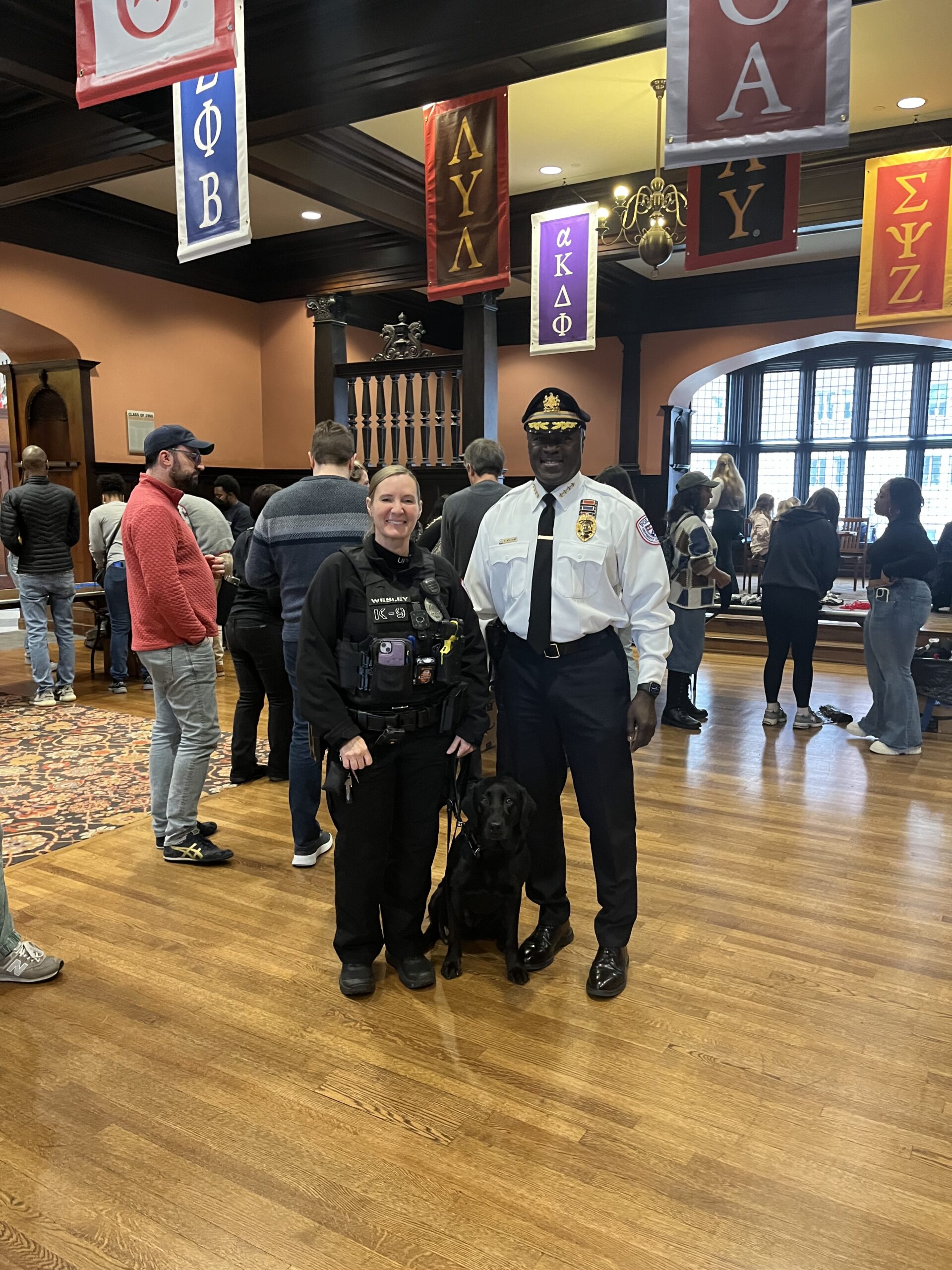 Chief Williams with Officer Wesley and K9 Officer Uman (a black labrador retriever) pose for a photo in Houston Hall as a volunteer event is taking place.
