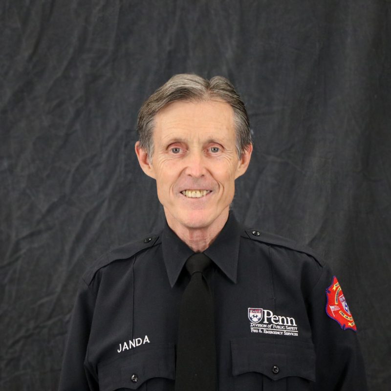 Photo of Gene Janda, with short black and gray hair, wearing a black shirt dress uniform shirt and black tie.