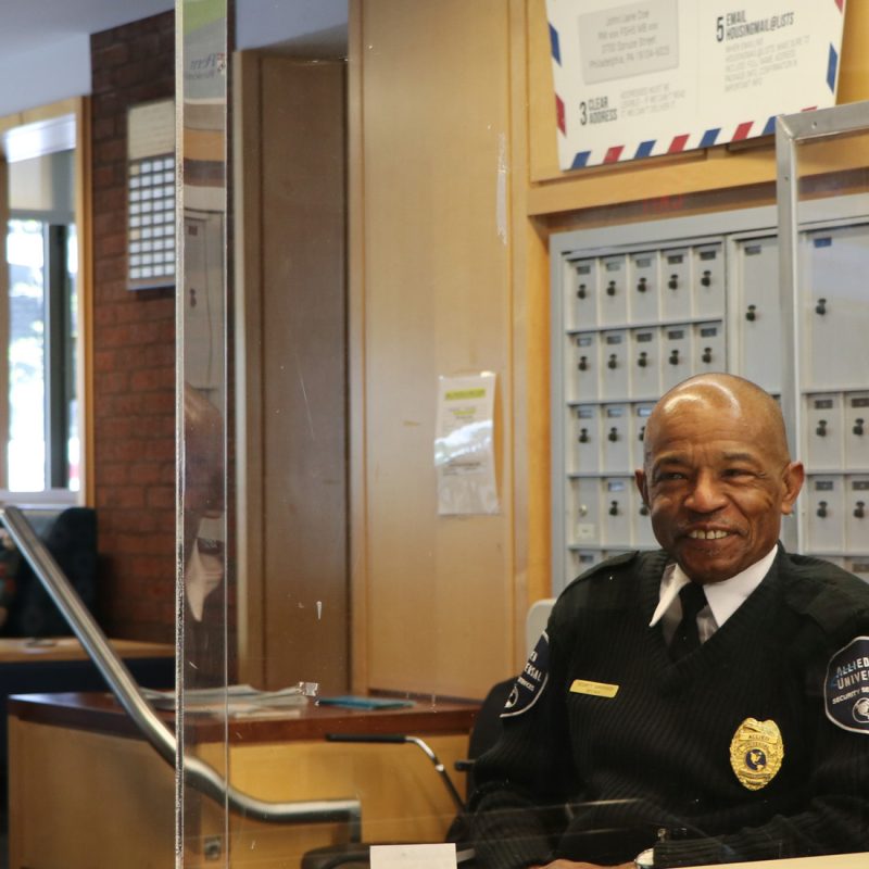 Security Officer wearing a black uniform sweater and badge sitting in the entrance to DuBois College House, smiling