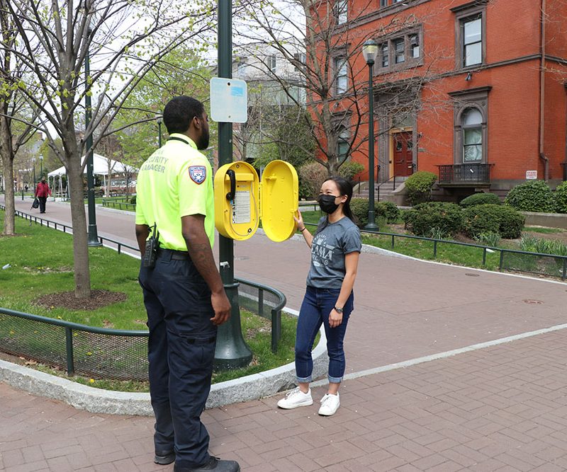 a student uses a Blue Light phone, and is greeted by a security officer wearing a bright yellow shirt.