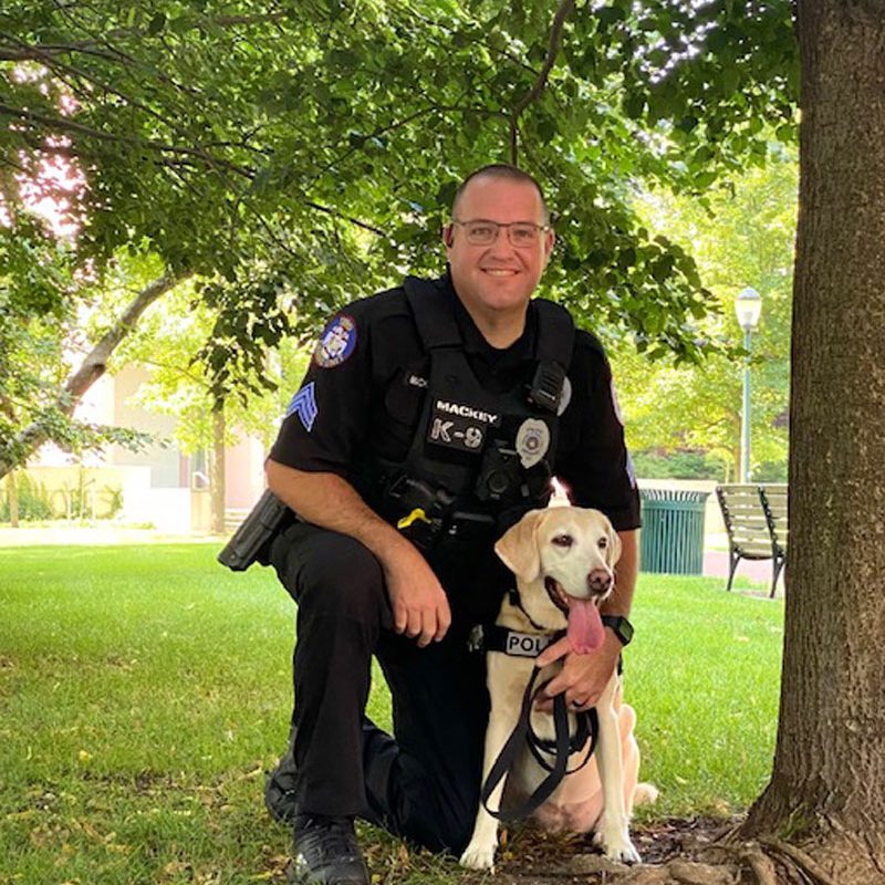 Sgt. Mackey, wearing a black police uniform, poses with canine Zzisa, a yellow labrador retriever, in a field under a tree.