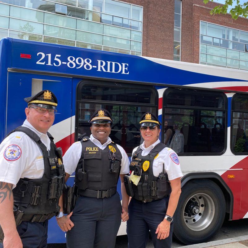 Lt. Bsuch, Capt. McCoy and Lt. VanDerVort pose in front of a Penn Transit bus, all wearing short sleeved-white dress shirst with black tactical vests.